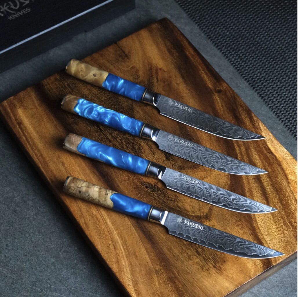 A set of Damascus knives with blue handles on a wooden cutting board.