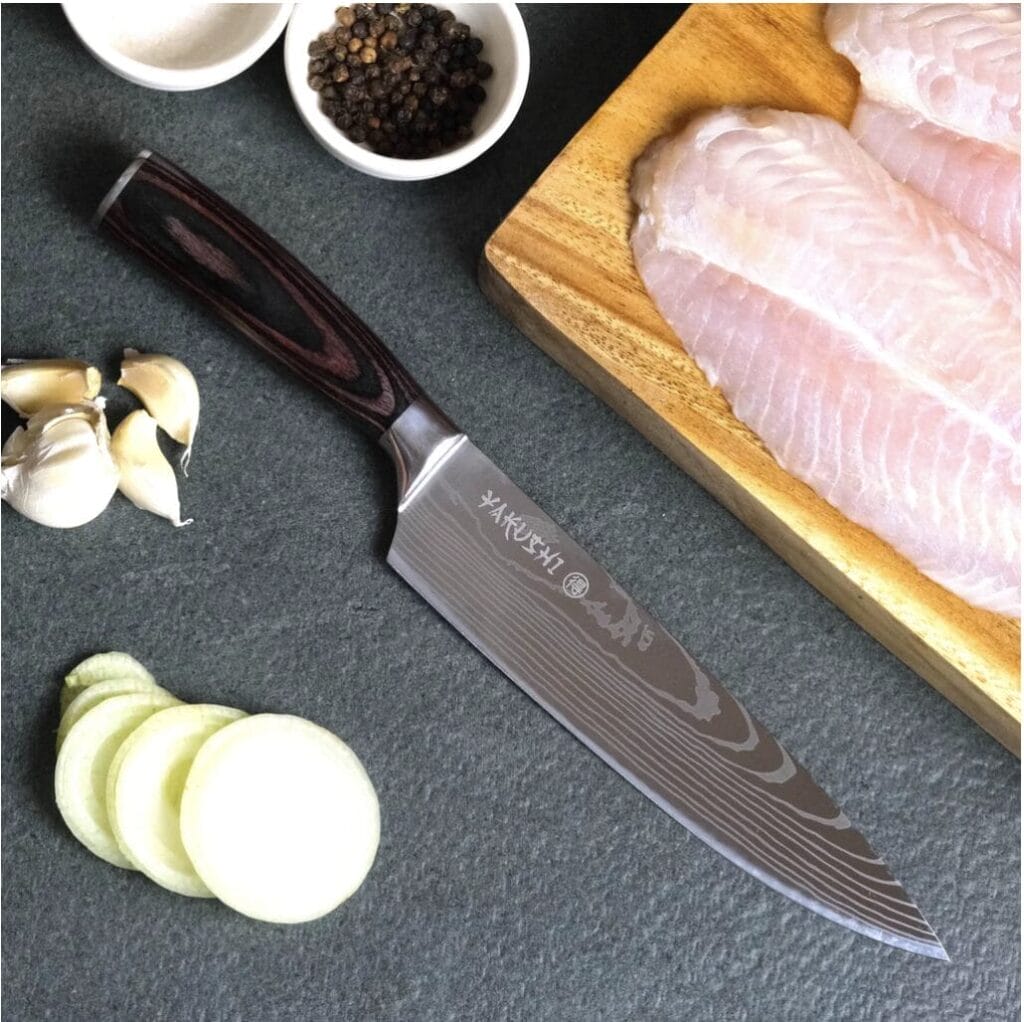 A Damascus santoku knife on a cutting board next to a piece of fish.